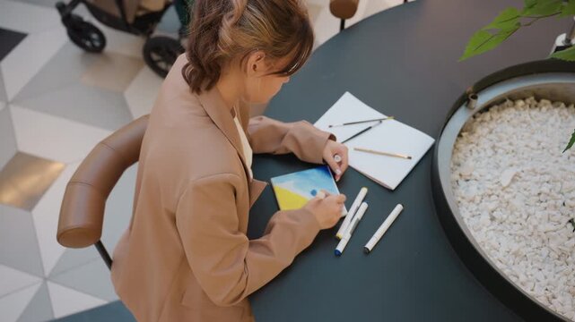 woman creating colorful artwork in open space, young female artist working carefully on detailed color illustrations, teenage woman engaged in precise drawing and coloring at round table in atrium