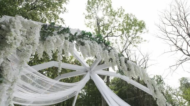 White wooden wedding arch decorated with hanging wisteria flowers and sheer fabric under green trees. Low angle shot