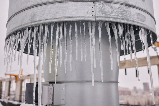 Metal vent with hanging icicles, Frozen icicles draping metallic chimney vent, Closeup of icy metal vent and chimney under overcast sky in winter storm conditions