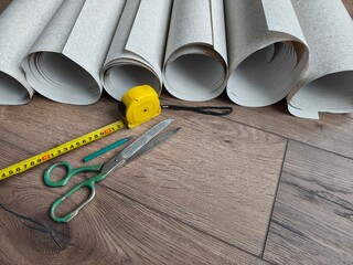 White wallpaper rolls with yellow measuring tape, green scissors, and a pencil on wooden floor for home renovation project.