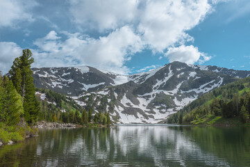 Fototapeta premium Scenic view to forest lake against large rocky mountain with white snow under lush clouds in blue sky. Reflection of snowy mountain on smooth water surface in sunlight. Coniferous trees on hilly shore