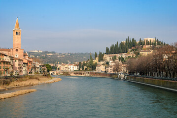 Fototapeta premium Scenic view of Verona along the Adige River with historic buildings, church tower and hillside skyline under a clear blue sky, capturing the romantic atmosphere of northern Italy.