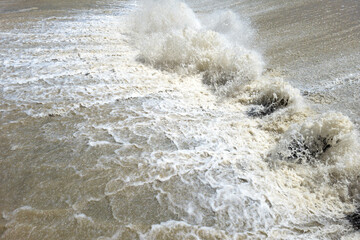 Powerful water splashes and churns vigorously in a natural outdoor setting. The scene captures the forceful movement of water under bright daylight, evoking a sense of energy and turbulence.