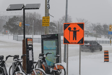 Obraz premium orange construction sign, Bike Share Toronto docking station and map at intersection of Lake Shore Blvd W and Ontario Drive during a snow storm on 2026-01-15
