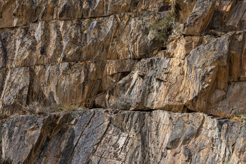 Rugged rock formation stands proudly in a mountainous region, surrounded by small resilient plants. The earthy tones and textures create a barren yet striking natural environment during daytime.