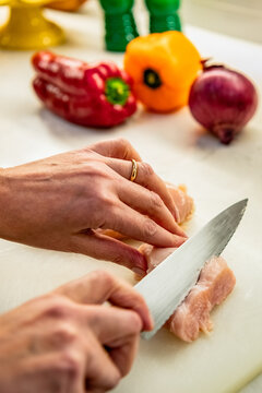A closeup photo showing the hands of a lady wearing a wedding band cutting a chicken in her kitchen with vegetables and utensils around the cutting board