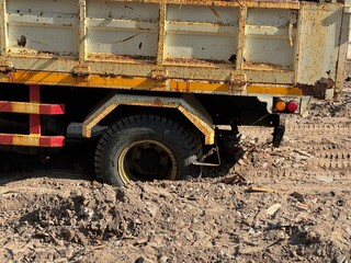 Rear wheel of an old rusty dump truck sunk into soft dirt at a construction site, showing heavy load impact, rough terrain, and ground instability.