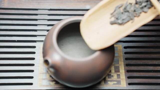 Top down slow motion of a traditional tea ritual on a tea board (chaban). Dry tea leaves are poured from a chahe into an empty brown clay teapot with careful hands.