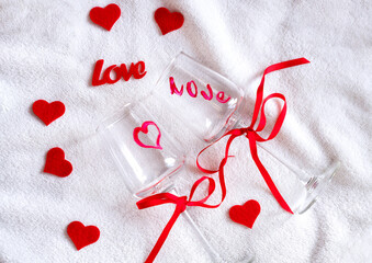 Cozy lifestyle Valentine's Day scene. Wine glasses painted with hearts and love word, red ribbon and felt hearts. Intimate home moment celebrating love and romance mood. Selective focus.