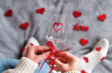 Woman's hands hold a wine glass decorated with a red ribbon and painted heart, surrounded by red hearts. Cozy Valentine's Day atmosphere, love and romance concept. Selective focus.