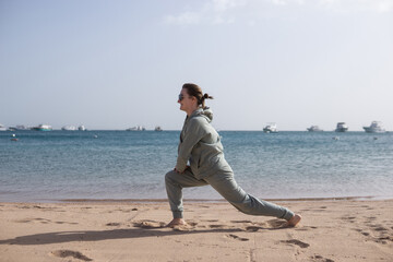 Woman in sports outfit doing lunge exercise on sandy beach with sea and boats background for outdoor fitness and healthy lifestyle concept.