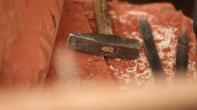 Close up view of a rusty hammer and chisels on red stone dust. Focus shifts to a wooden measuring tool held by a sculptor in the foreground.

