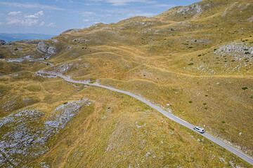 Aerial view of a camper van driving along a winding road at Sedlo Pass in Durmitor, Montenegro, surrounded by rugged mountain landscapes, capturing freedom, adventure, and scenic road travel.