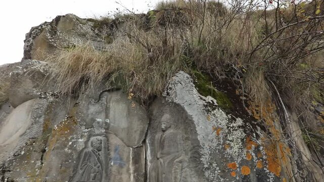 A rock wall featuring carved Soviet-era memorials. The shot moves past a carved USSR emblem to reveal a larger bas-relief of historical figures carved directly into the natural cliff face.

