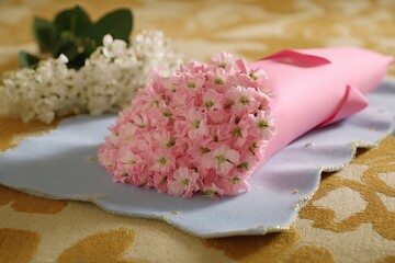 A bouquet of pink flowers wrapped in paper rests on a blue cloth with white flowers placed beside it in a bright setting