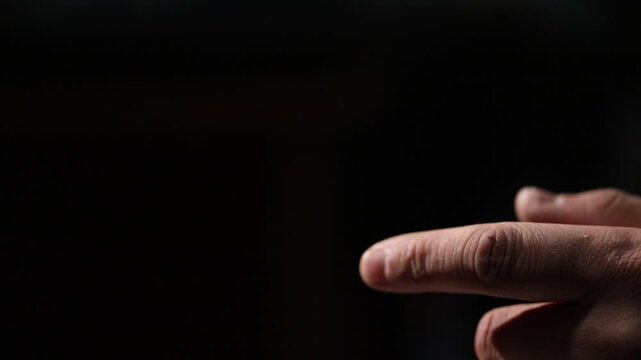 Close up shot of male hands interlocked on knee in dim red lighting. The man moves his fingers and unclasps his hands, making a small gesture in darkness.


