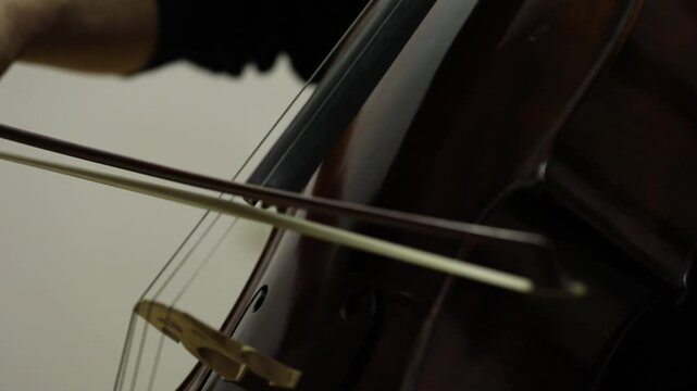 Close up shot of a musician playing the cello. The cellist wears a white shirt and uses a bow on the strings of the classical wooden instrument.

