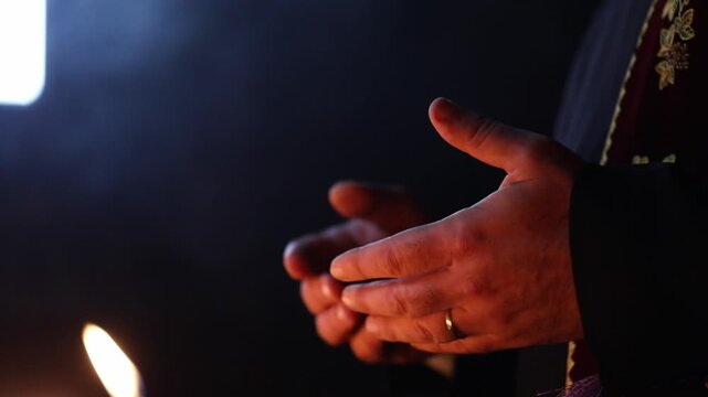 Close up side view of the hands of a priest wearing traditional vestments making the sign of the cross during a religious ceremony with a dark background.

