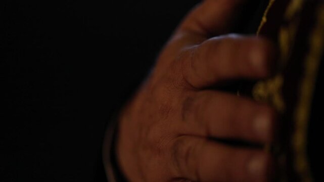 Extreme close-up of a priest's hand resting on a dark red vestment with gold embroidery, conveying deep faith and reverence during a quiet moment of prayer.


