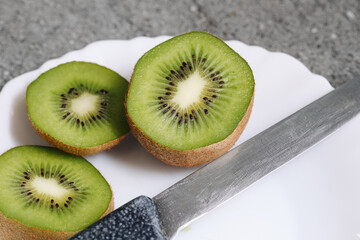 Fresh kiwi fruit sliced in halves on white plate. Sharp kitchen knife next to green kiwi closeup. Healthy food preparation scene. Natural vitamin rich fruit ready for eating at home.