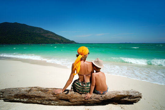 Rear view of a mother and child sitting together on a sandy tropical beach and looking at the turquoise sea. Summer vacation, family travel and lifestyle concept. 