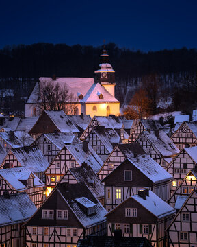 Snow covers houses in a small town under a night sky near a church with a clock tower