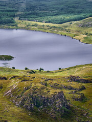 A lake stretches out surrounded by green hills and trees. Rocks form a terrace in the foreground. The day appears cloudy.