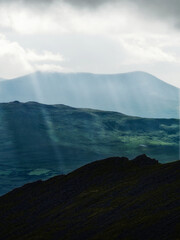 Sun rays filter through the cloudy sky above rolling green hills and a dark mountain ridge. The scenery shows a tranquil Irish landscape.