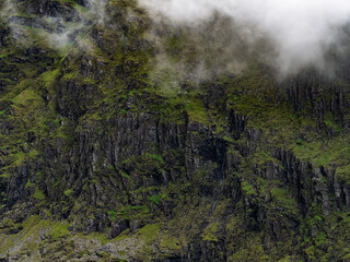 A detailed view of a mountain face. Dark rocks are covered with patches of green moss and vegetation. Low lying clouds and mist cover the upper portion of the slope.