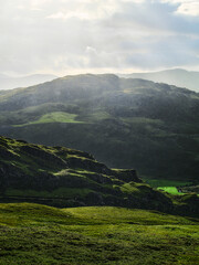 A scenic view with sunlight hitting the green hills and mountains. The overcast sky creates a peaceful atmosphere over the vast landscape.