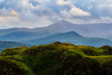 Green hills sit in the foreground, leading back to blue layered mountains. Clouds cover the sky above National Park during the daytime.