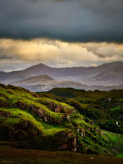 A green, vegetated hillside with rocky outcrops slopes down into a valley. In the background, distant mountains rise under a cloudy sky in the national park.
