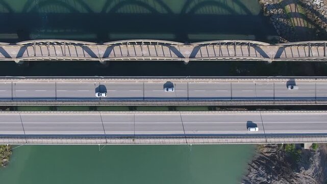 Aerial view shows old concrete arch bridge beside new highway bridge crossing river. Parallel transport structures highlight historic and modern engineering across flowing water.
