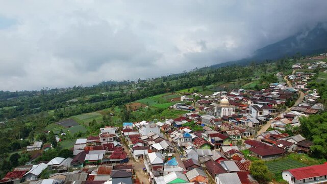 Aerial drone footage of a local village, in an agricultural valley of vegetables, with mountain slope, dark cloudy atmosphere, in Tegal regency, nearby Slamet volcano, Java island, Indonesia