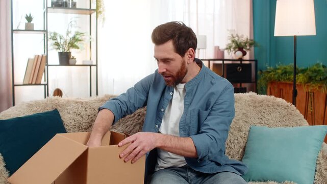 Young man at home unpacks a package, eagerly pulling out a colorful shirt and admiring it with joy. Caucasian guy smiles while holding the new clothing, excited about his online shopping purchase