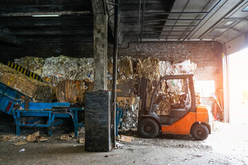 An orange forklift is in a warehouse environment. It appears to be idle next to large bales of paper and cardboard intended for recycling
