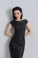 Elegant brunette woman with red lipstick in a black patterned dress leaning against a gray wall.