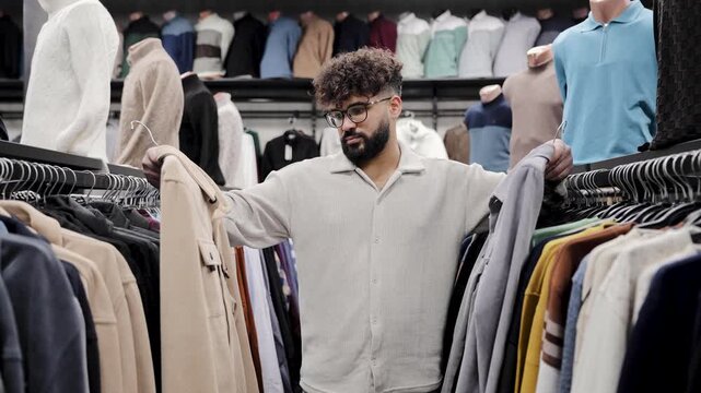 Young man standing in clothing store, holding two garments on hangers, thinking about purchase for his wardrobe
