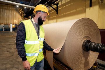 Man worker in safety gear inspects check massive rolls of paper inside a manufacturing facility. The scene captures the busy atmosphere of the production process during daylight hours.