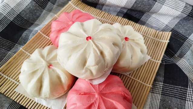 Steamed buns known as baozi or bakpao served on bamboo mat over black and white checkered cloth. Soft pleated buns with red dot detail in white and pink colors.