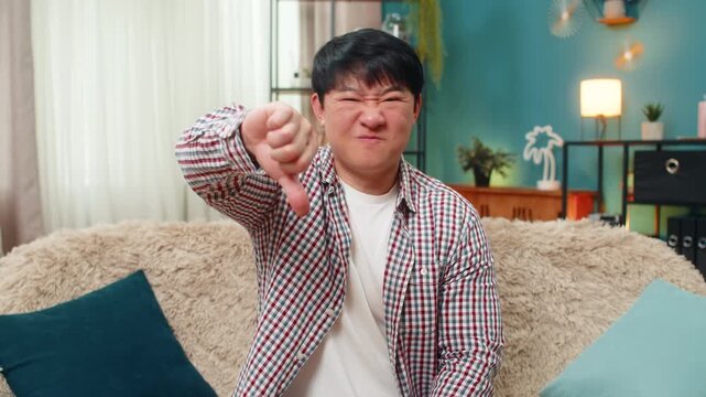 Asian man shows thumbs down gesture frowning unhappily to express negative disapproval. Chinese guy sits on home sofa shows strong disagreement signaling rejection with stern expression clear feedback