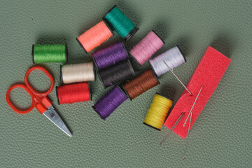 Spools thread in different colors, scissors and sewing needles on green leather background.