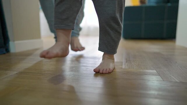 Toddler boy walks barefoot near adult leg on home floor. Child takes slow step while learning to move balance. Boy practices balance carefully. Child focuses on balance with each small walk indoors.