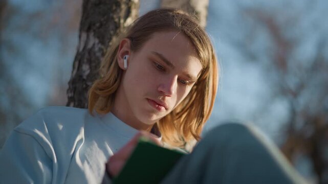 youthful author reflecting, young novelist jotting down inspirations amidst tranquil park scenery, creative individual capturing reflective insights while seated comfortably under lush tree branches