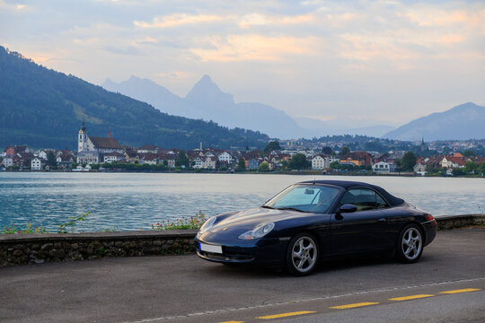 Arth, Switzerland - July 5, 2025: Blue Porsche 911 convertible next to Lake Zug (Zugersee) with village Arth and mountains in canton of Schwyz in Switzerland