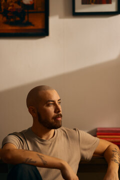 A contemplative man with a beard sits in a warm, indoor setting with dramatic lighting. His thoughtful expression and visible tattoos create a moody, authentic portrait of modern masculinity.