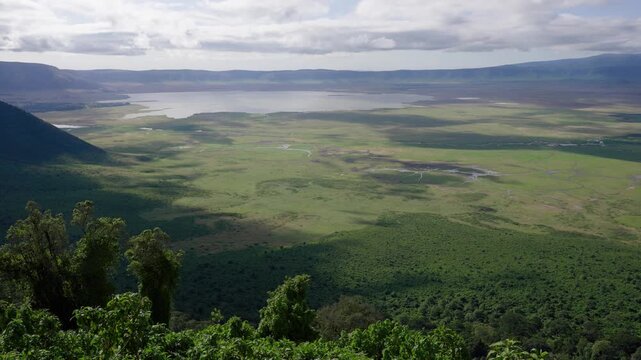 Wide panoramic view of Ngorongoro Crater caldera with sweeping grasslands, wetlands, and crater rim under moving clouds, filmed in slow panning motion.