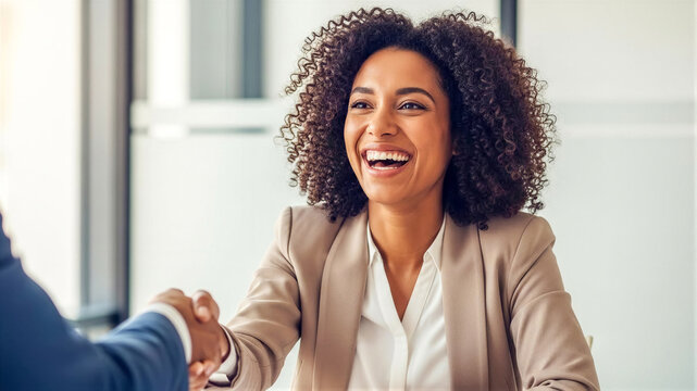 Smiling businesswoman with voluminous curly hair in gray suit warmly shaking hands with another person across a desk in bright modern office, suggesting successful interview or deal