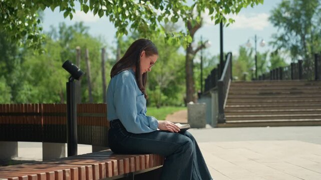 young caucasian woman reading book on bench near stairs, reflective traveler or student enjoying guidebook in urban park, leafy canopy and concrete steps, relaxed pose with soft midday light