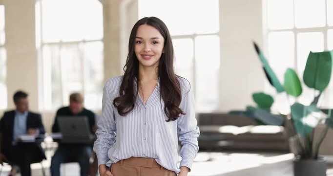 Professional female office employee posing, looking at camera standing in corporate workspace, business colleagues in background. Company staff member or applicant portrait, career, human resources